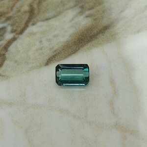 Op de afbeelding: Een groene en blauwe smaragd geslepen edelsteen op een wit marmeren oppervlak.