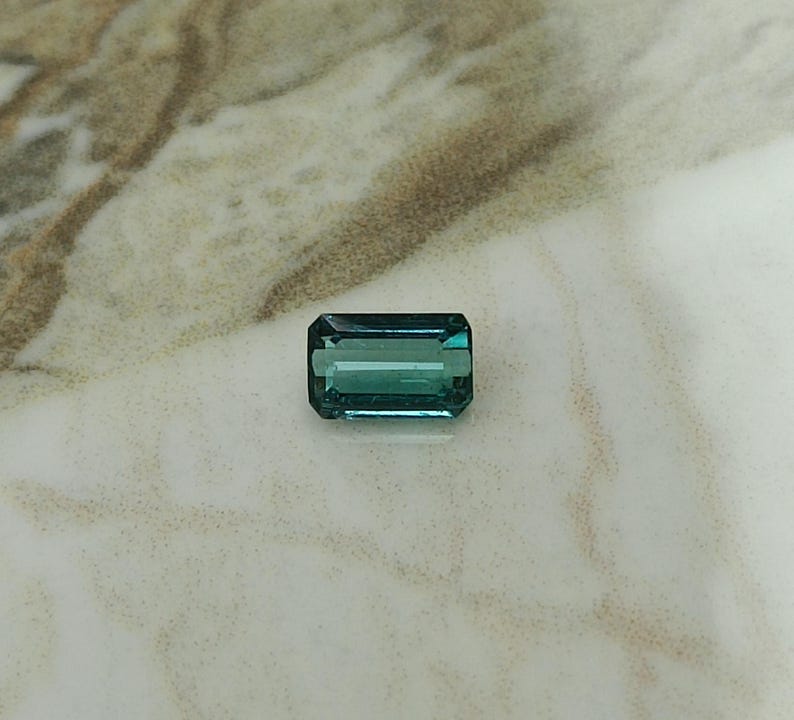 Op de afbeelding: Een groene en blauwe smaragd geslepen edelsteen op een wit marmeren oppervlak.