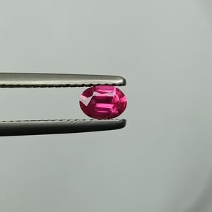 Puede incluir: Una vibrante gema rosa intenso, de corte ovalado, es sostenida por unas pinzas. Las facetas de la gema reflejan la luz, creando un efecto brillante. El fondo es un gris suave y neutro.