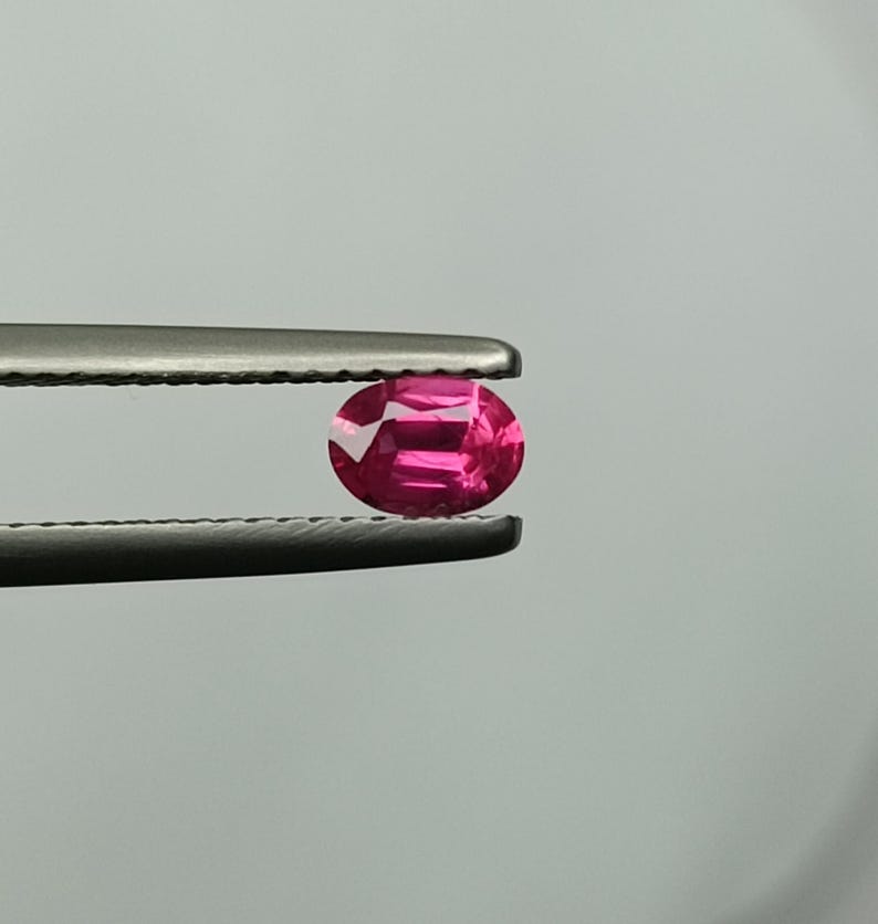 Puede incluir: Una vibrante gema rosa intenso, de corte ovalado, es sostenida por unas pinzas. Las facetas de la gema reflejan la luz, creando un efecto brillante. El fondo es un gris suave y neutro.