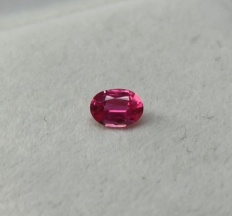 Puede incluir: Una gema rosa, de talla ovalada. La gema tiene una superficie pulida con facetas internas que reflejan la luz. El color es un rosa intenso y saturado, sobre un fondo neutro. Esta piedra preciosa es adecuada para joyer&iacute;a.