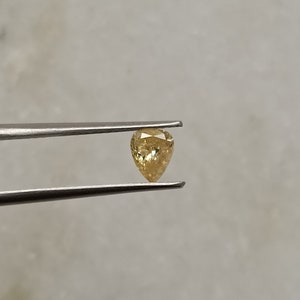 May include: A pear-shaped, yellow gemstone with a faceted cut. The gemstone is held in tweezers against a white background.