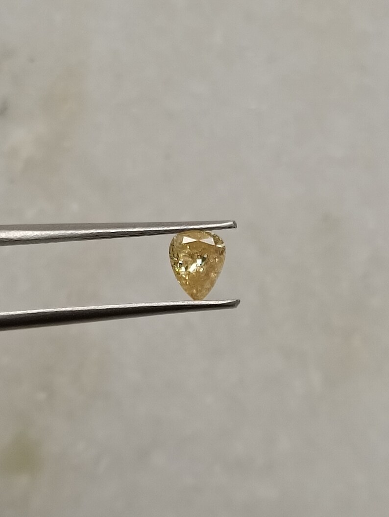 May include: A pear-shaped, yellow gemstone with a faceted cut. The gemstone is held in tweezers against a white background.