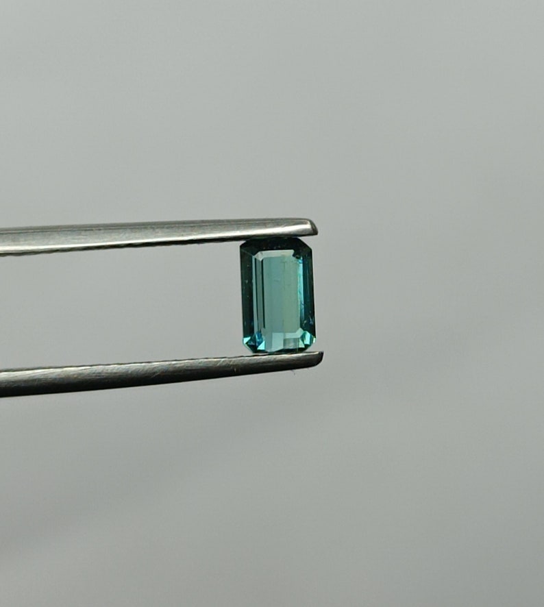 Op de afbeelding: Een teal blauwe edelsteen met een rechthoekige smaragd-slijp, vastgehouden in een pincet.
