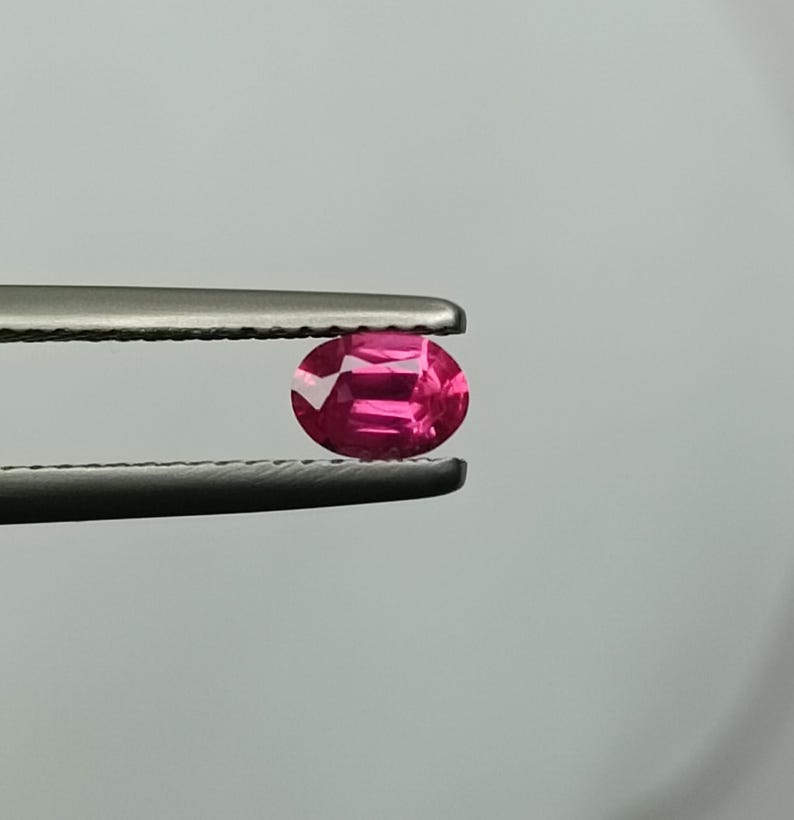 Puede incluir: Una vibrante gema rosa, de corte ovalado, es sostenida por unas pinzas plateadas sobre un fondo neutro. Las facetas de la gema reflejan la luz, mostrando su claridad y color. Esta imagen resalta el brillo de la gema.