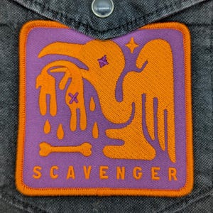 Puede incluir: Un parche cuadrado con un diseño naranja y morado, que presenta una figura de pájaro sosteniendo una figura pequeña. La palabra "SCAVENGER" está impresa en blanco en la parte inferior. El parche está cosido en un bolsillo de camisa vaquera gris.