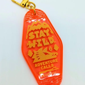 May include: Orange motel-style keychain with gold text and hardware. The keychain reads "Stay Wild, Adventure Calls" with mountain, leaf, and animal graphics. The keychain is made of a clear, orange acrylic material.