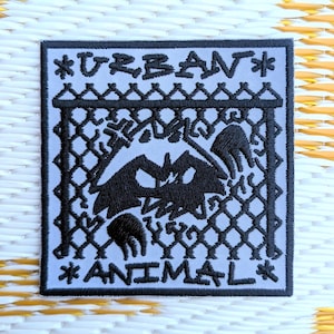 Könnte beinhalten: Quadratischer Aufnäher mit hellblauem Hintergrund und schwarzem Stickdesign. Der Aufnäher zeigt einen stilisierten Waschbären oder ein ähnliches Tier hinter einem Zaun, mit den Worten "URBAN" und "ANIMAL" über und unter dem Bild.