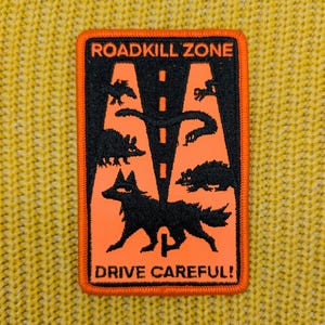 May include: A rectangular patch with an orange border. The patch has a black and orange design featuring the text "ROADKILL ZONE" and "DRIVE CAREFUL!". The design includes a road with animal silhouettes.