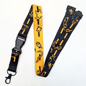 May include: A fabric lanyard featuring a repeating pattern of yellow animal silhouettes and road markings on a black background. The strap includes a black plastic side-release buckle and a black metal swivel lobster clasp at the bottom end.