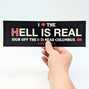 May include: Black bumper sticker with the text "I ❤️ THE HELL IS REAL" in white and red lettering. The text "SIGN OFF THE I-71 NEAR COLUMBUS, OH" is also included. The bottom of the sticker reads "MOT 77".
