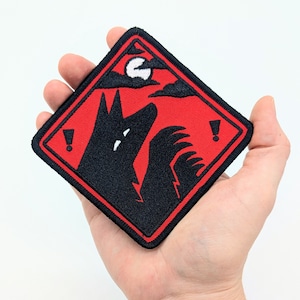 Pu&ograve; includere: Toppa quadrata con una silhouette nera di lupo che ulula alla luna piena su sfondo rosso. La toppa ha un bordo nero e due punti esclamativi. Il design ricorda un segnale di avvertimento.