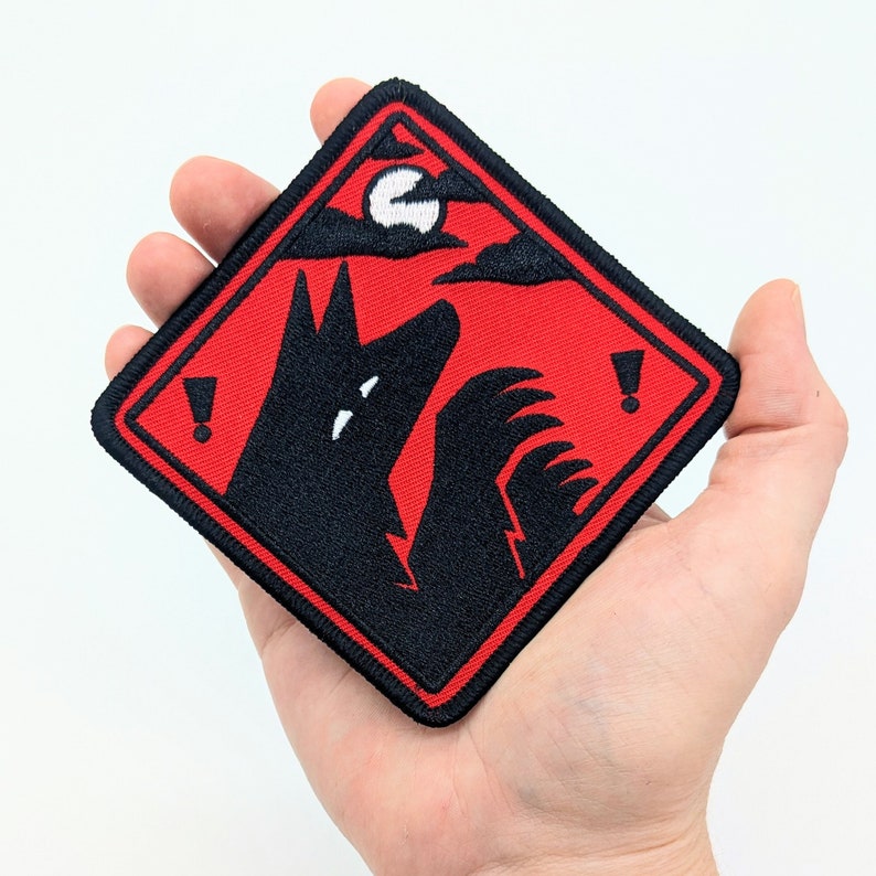 Pu&ograve; includere: Toppa quadrata con una silhouette nera di lupo che ulula alla luna piena su sfondo rosso. La toppa ha un bordo nero e due punti esclamativi. Il design ricorda un segnale di avvertimento.