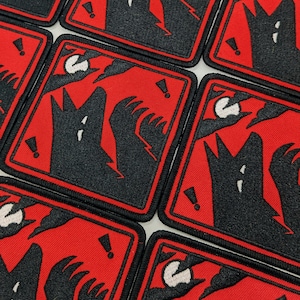 Könnte beinhalten: Quadratische, gestickte Aufnäher mit einem schwarzen Wolf, der den Mond anheult, auf rotem Hintergrund. Das Design umfasst Blitze, Ausrufezeichen und einen schwarzen Rand. Die Aufnäher sind wahrscheinlich für Kleidung oder Accessoires.