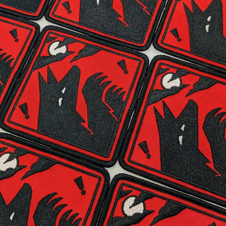 Pu&ograve; includere: Toppe quadrate ricamate con un lupo nero che ulula alla luna su sfondo rosso. Il design include fulmini, punti esclamativi e un bordo nero. Le toppe sono probabilmente per abbigliamento o accessori.