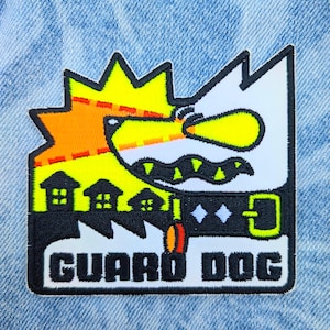 May include: A rectangular patch featuring a cartoon dog design with the text "GUARD DOG". The patch has a white dog head with a yellow and orange background, a black collar, and a small house graphic.