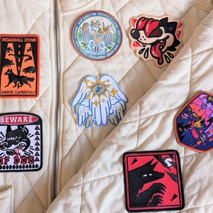 Pu&ograve; includere: Collezione di toppe ricamate con vari disegni, tra cui un lupo, ali d'angelo e un cane dei cartoni animati. Le toppe presentano colori vivaci come rosso, blu e arancione, e alcune includono testo come "ROADKILL ZONE" e "BEWARE".