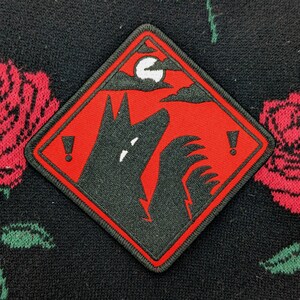 Pu&ograve; includere: Toppa ricamata nera e rossa a forma di diamante. Raffigura un lupo che ulula alla luna, con sfondo rosso e bordo nero. Due punti esclamativi affiancano l'immagine del lupo.