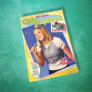 May include: A book cover for "Club Stephanie: Forget It, Flamingoes!" based on the TV series "Full House". The cover features a young woman holding a microphone and wearing a blue crocheted top with a flower design. The text "Forget It, Flamingoes!" is printed on the cover.