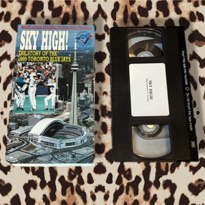 Może przedstawiać: Wyświetlono kasetę VHS i jej okładkę. Okładka zawiera tytuł "SKY HIGH!" z podtytułem "THE STORY OF THE 1989 TORONTO BLUE JAYS". Taśma ma etykietę z napisem "SKY HIGH" i "Approximately 55 minutes."