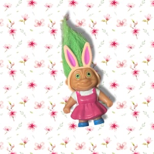 May include: A small troll figurine with bright green hair and pink bunny ears. The figure has a tan complexion, green eyes, and a wide smile. It is wearing a white short-sleeved shirt under a solid pink dress and small blue shoes on its feet.