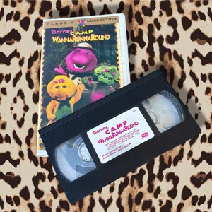 Può includere: Una videocassetta VHS e la sua custodia per "Barney Camp WannaRunaround" con Barney il dinosauro e i suoi amici. La custodia presenta un'immagine colorata dei personaggi. La videocassetta VHS nera è in primo piano.