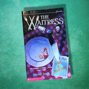 May include: A paperback book titled The Waitress by Sinclair Smith, featuring a cover illustration of a plate with food remnants, a knife, forks, a salt shaker, and a restaurant check that reads Have a Nice Day on a purple checkered tablecloth.