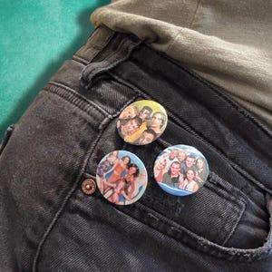 May include: Three circular pin-back buttons attached to the pocket of black denim jeans. Each button features a glossy, full-color photographic print of a group of people. The buttons are arranged in a cluster on the dark fabric of the jeans.