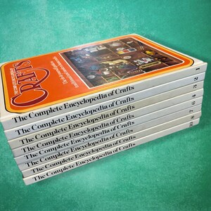 May include: A stack of white books titled "The Complete Encyclopedia of Crafts." The top book has an orange cover with the word "Crafts" in large, stylized letters. The books are stacked on a teal surface.