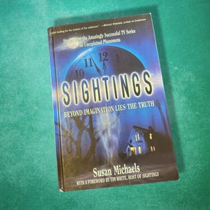 Op de afbeelding: Een boekomslag met een donkerblauwe achtergrond en een klok met het woord "SIGHTINGS" in witte letters. De titel van het boek is "SIGHTINGS: BEYOND IMAGINATION LIES THE TRUTH". De auteur is Susan Michaels. Het boek is gebaseerd op de tv-serie over onverklaarbare fenomenen.