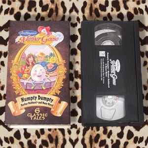 May include: A VHS tape and its case for "Jim Henson's Mother Goose Stories: Humpty Dumpty & Many More." The case is brown with a gold frame and features illustrations of characters. The VHS tape is black.