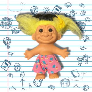 May include: A troll doll with bright yellow hair and a black graduation cap featuring a yellow tassel. The doll has a peach-colored skin tone and is wearing pink shorts decorated with a repeating pattern of small blue palm trees and yellow suns.