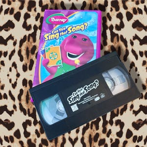 Può includere: Una cassetta VHS e custodia viola di Barney con il titolo "Can You Sing that Song?". La custodia presenta Barney che tiene delle carte. La cassetta VHS nera è etichettata con lo stesso titolo.