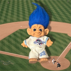 May include: A troll doll with bright blue hair, wearing a cream-coloured baseball uniform. The shirt features a 1992 World Series logo, and the trousers are printed with the number 32 in blue. The doll has tan skin, large ears, and a wide smile.