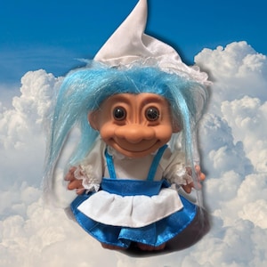 May include: A vintage troll doll with blue hair, wearing a white bonnet and a blue and white dress. The doll has a smiling expression and large, round eyes. The background is a blue sky with white clouds.