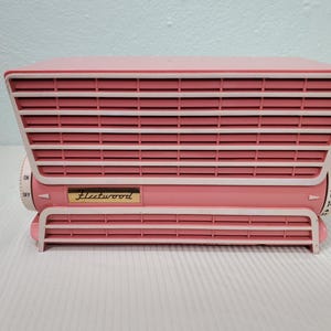 Könnte beinhalten: Ein rosa-weißes elektrisches Retro-Ventilator. Der Ventilator hat eine rechteckige Form mit horizontalen Lamellen und abgerundeten Kanten. Der Name "Fleetwood" ist auf der Vorderseite zu sehen. Er hat ein Zifferblatt mit Zahlen.