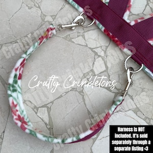 May include: Close-up of a pet harness with a floral pattern in red, green, and white on a white background. The harness has a burgundy strap and silver metal clasps. The text "Crafty Crinkletons" is visible.