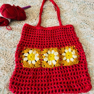 May include: A handmade red crochet tote bag with a rectangular floral design in yellow and white. The bag has a mesh-like pattern and two red handles. Red yarn and a crochet hook are visible.