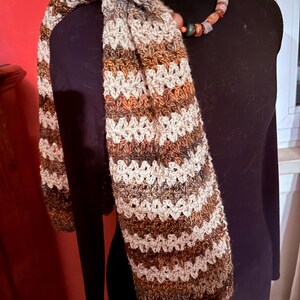 May include: A handmade, striped scarf in shades of brown, white, and orange. The scarf is draped over a black form, showcasing its textured, crocheted design. A beaded necklace with earth-tone beads is also visible.