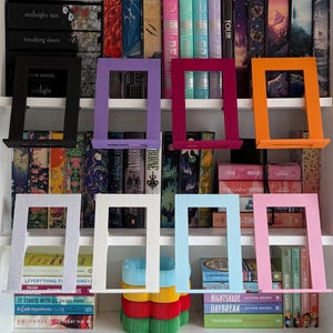 Op de afbeelding: Acht rechthoekige metalen boekenstandaards in diverse kleuren, waaronder zwart, paars, magenta, oranje, zilver, wit, lichtblauw en roze, gerangschikt in twee rijen voor een witte boekenkast gevuld met talloze boeken van verschillende formaten.