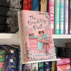 Peut inclure: Un livre intitulé The Break Up Pact par Emma Lord, présenté sur un support d'étagère blanc. La couverture illustre deux personnes assises à une table, entourées de motifs floraux, avec un sol à carreaux roses et blancs en arrière-plan.
