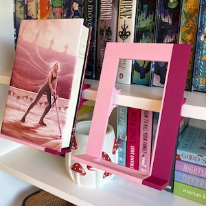 Peut inclure: Un support de livre bicolore rose et magenta tenant un livre avec une illustration de couverture représentant une personne tenant une épée. Le support présente un design à cadre rectangulaire avec un rebord inférieur pour soutenir le livre.