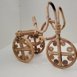 May include: A small, decorative tricycle made of woven rattan. The tricycle is brown and features a unique design with circular details on the wheels.