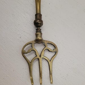 Könnte beinhalten: Ein Vintage-Kaminwerkzeug aus Messing mit einem dekorativen Griff und einem dreizackigen Gabeldesign. Der Griff hat ein gedrehtes Muster und die Gabel ein durchbrochenes Design. Das Werkzeug wird wahrscheinlich zum Anzünden eines Feuers verwendet.