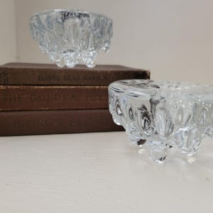 May include: Two clear glass candle holders with a textured, bubble-like design. The candle holders are resting on a stack of three vintage books with brown covers. The books have titles printed on the spines.