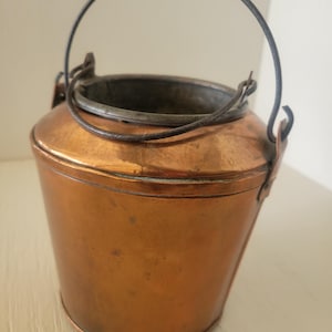 May include: A small, antique copper bucket with a dark metal handle. The bucket has a rounded body and a slightly flared rim. The handle is attached to the sides of the bucket with metal loops. The copper has a warm, reddish-brown color.