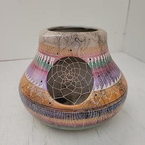 Puede incluir: Un jarrón decorativo de cerámica con un diseño único. El jarrón tiene forma redonda con una abertura ancha y un exterior colorido y estampado. El diseño incluye un elemento atrapasueños y una variedad de colores, incluyendo naranja, morado y rosa.