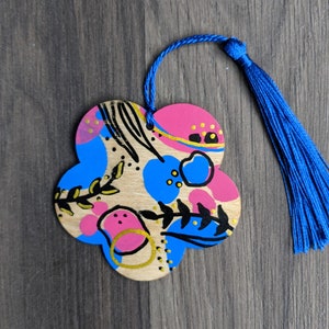 May include: A wooden, flower-shaped ornament with a blue tassel. The ornament is painted with pink, blue, and gold abstract shapes and black line art. A blue cord is attached for hanging.