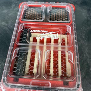 May include: A red and clear Milwaukee Packout storage container with multiple compartments. The container has a clear lid, allowing visibility of the contents. The Milwaukee logo is visible on the front. The container is designed for impact resistance and is IP65 rated.