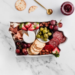 Könnte beinhalten: Ein weißes Keramik-Serviertablett in Form des Bundesstaates Washington, gefüllt mit einer Auswahl an Snacks, darunter Cracker, Käse, Trauben, Salami, Erdbeeren, Mandeln und Oliven.